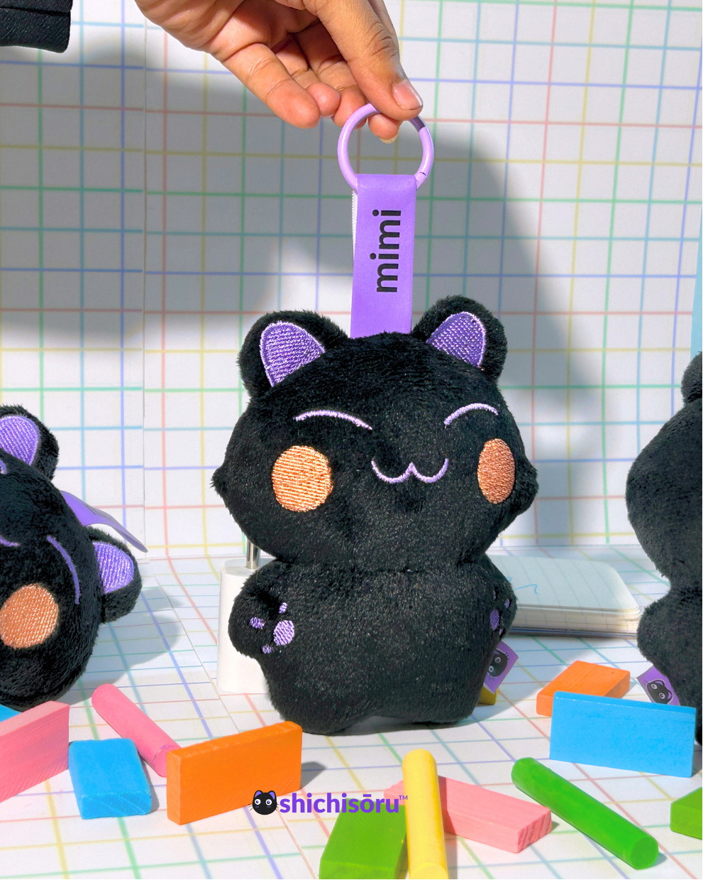 Black cat-shaped plush toy with purple accents held by a hand against a grid background.